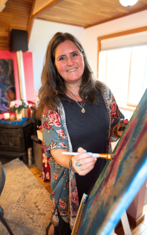Intentional Creativity facilitator Erika Primozich of Boulder, CO miles into the camera while holding a paintbrush next to a canvas during a painting workshop for self-exploration